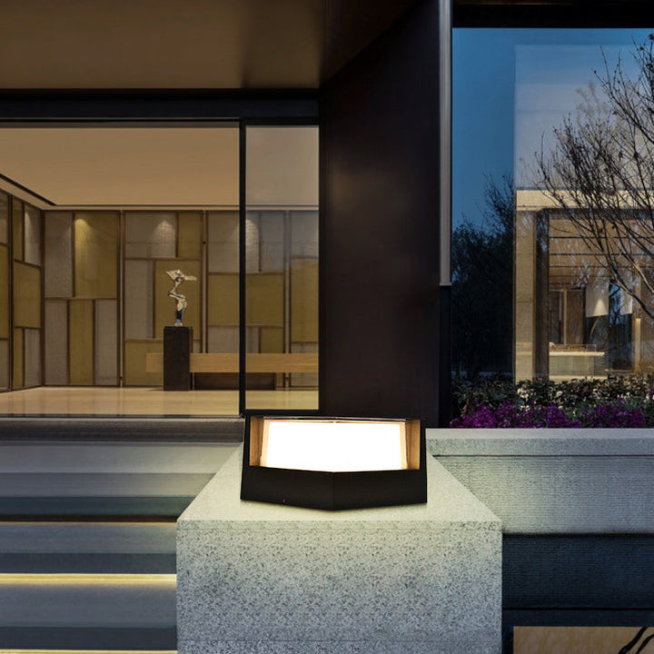 Fünfeckige schwarze Außenleuchte mit warmem Licht auf einer Steinmauer vor einem modernen Gebäude mit Glasfront und beleuchteten Stufen. 3