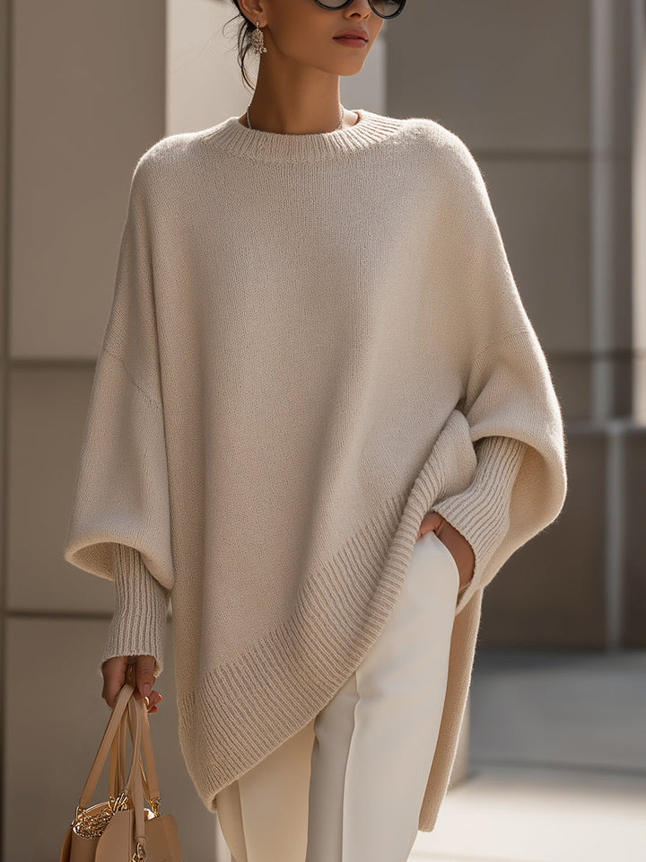 Beiger Strickponcho mit gerippten Bündchen, getragen mit weißer Hose und Handtasche vor einer hellen Steinwand 2