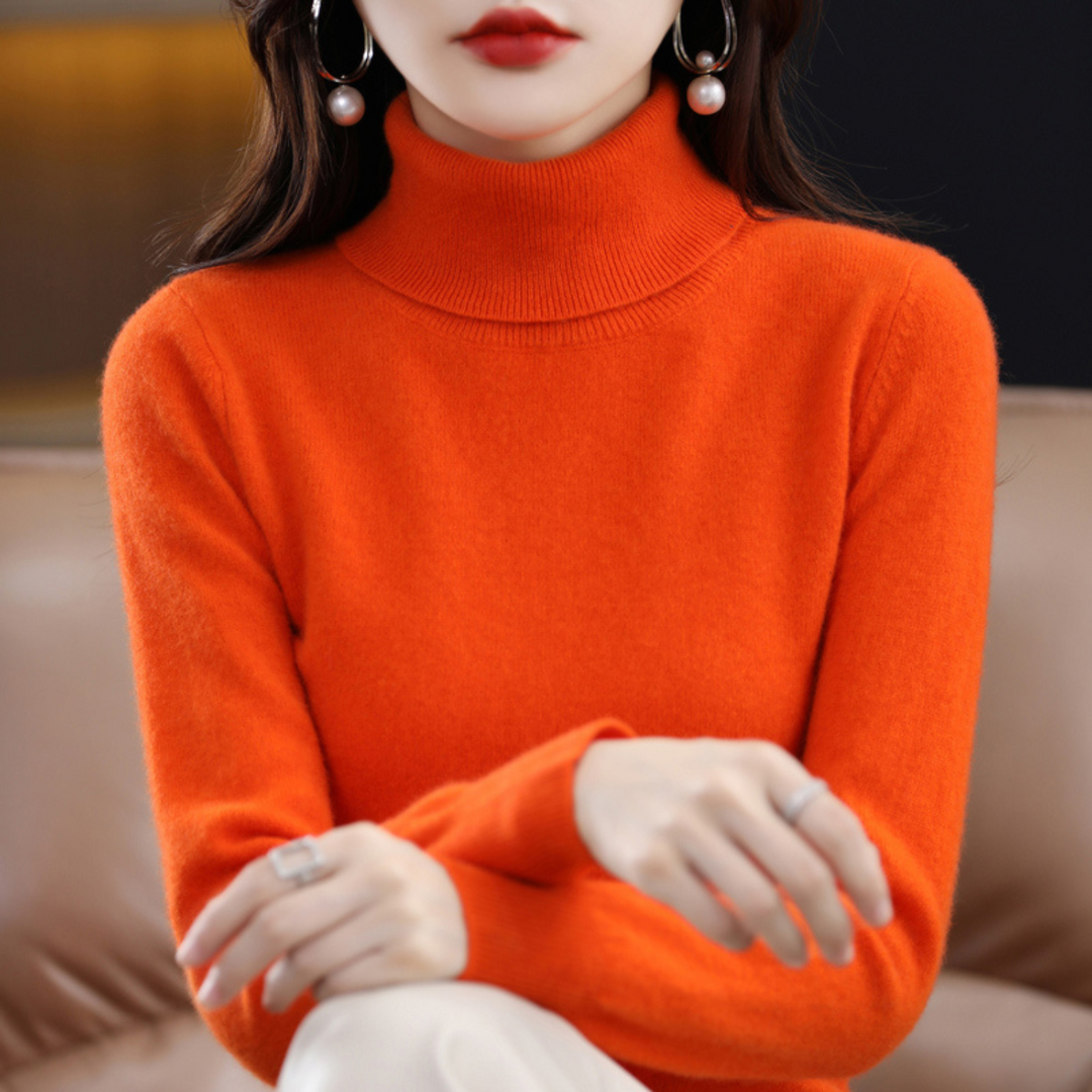 Oranger Rollkragenpullover am sitzenden Model mit überkreuzten Händen, dazu eine weiße Hose. 10