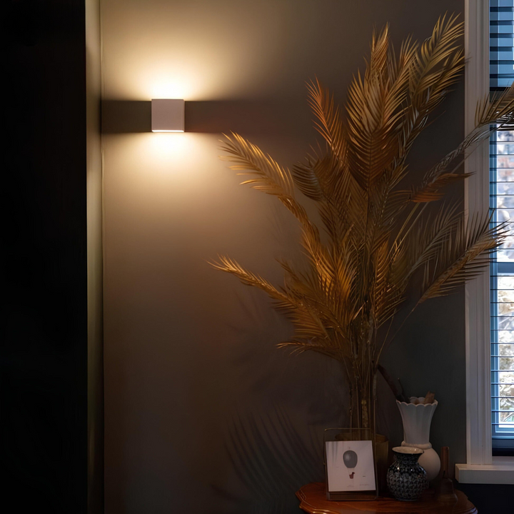 Weiße Würfelleuchte an einer dunklen Wand neben einer Vase mit goldenen Palmwedeln und einem Fenster, warmes Licht nach oben. 4