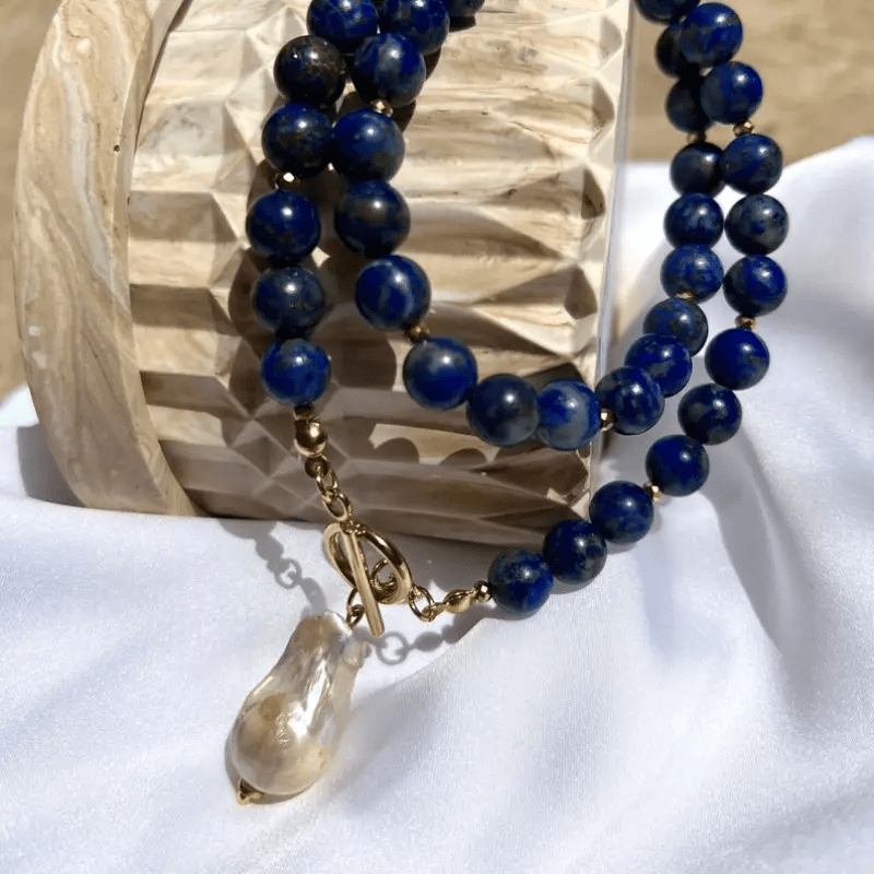 Doppelt gelegte Kette aus blauen Perlen auf weißem Stoff vor einem hellen, facettierten Holzobjekt 7