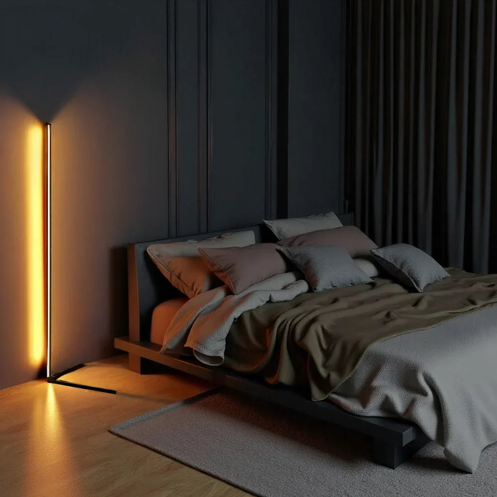 Stehlampe strahlt orange-gelbes Licht an dunkler Wand neben einem niedrigen Bett mit grauen Kissen 2