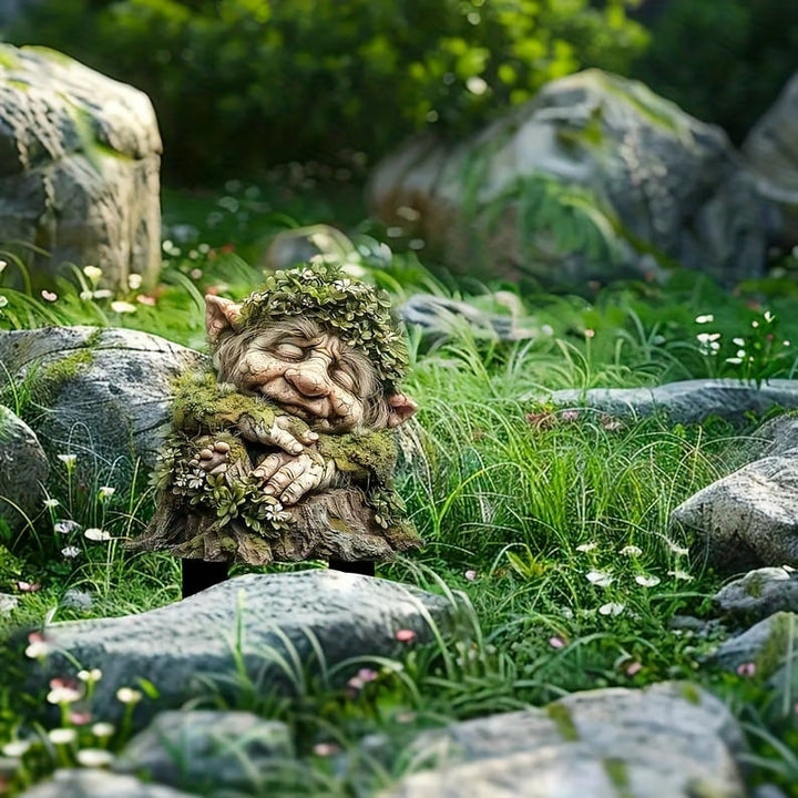 Schlafender Troll mit Blätterkranz und verschränkten Händen auf Baumstumpf, zwischen Felsen im Gras mit kleinen weißen Blüten. 16