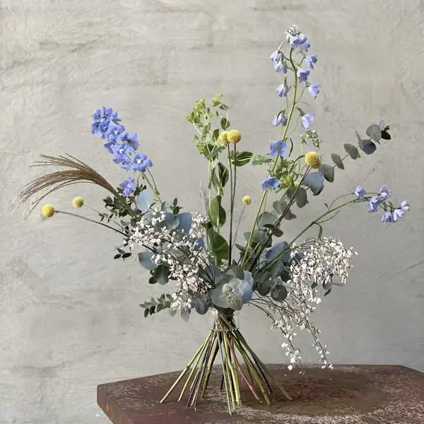 Gebundener Strauß mit blauem Rittersporn, gelben Kugelblumen und Eukalyptus auf rostigem Metalltisch vor grauer Wand. 12