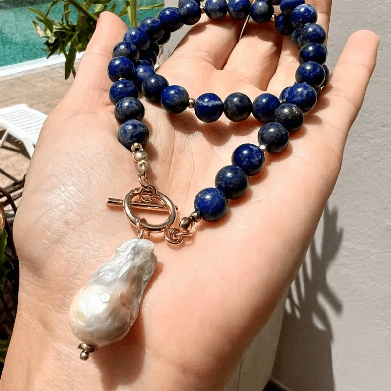 Blaue Perlenkette mit großer Barockperle liegt auf einer geöffneten Handfläche, im Hintergrund ein Pool 2