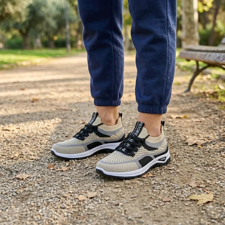 Beige-schwarze Sneaker an den Füßen einer Person im Park, dunkelblaue Jogginghose, Kiesweg mit Herbstlaub und Bank. 38
