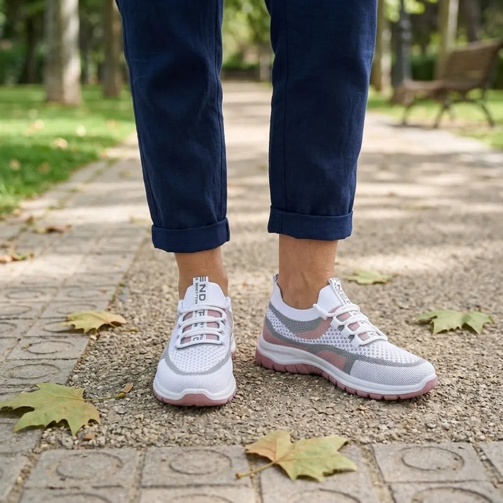 Weiß-rosa Mesh-Sneaker an den Füßen einer Trägerin mit dunkelblauer Hose auf einem Parkweg mit Herbstlaub. 20