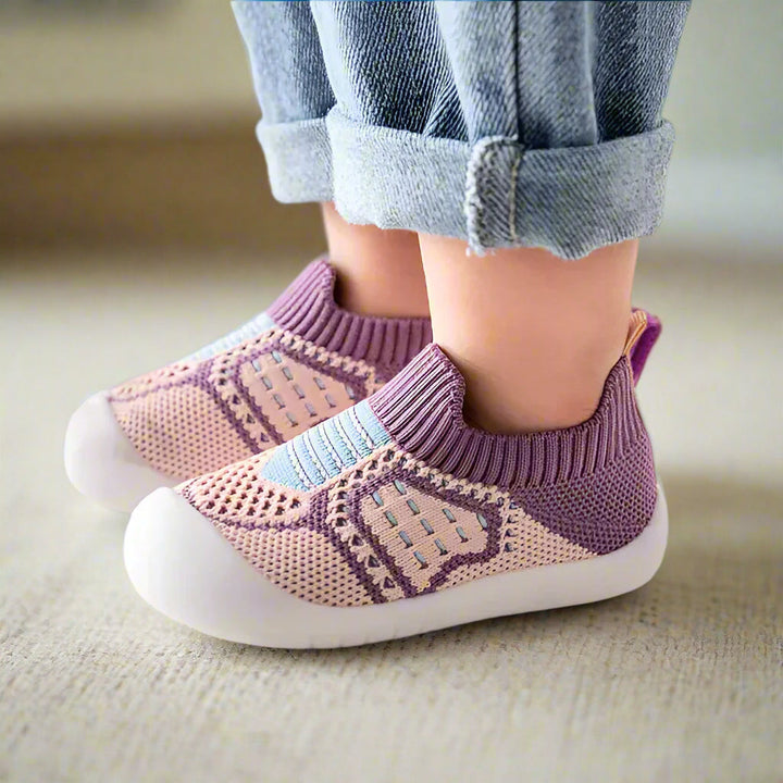 Kinderfüße in lila-rosafarbenen Strickschuhen mit hellblauen Streifen und weißer Gummisohle auf hellem Teppich. 15