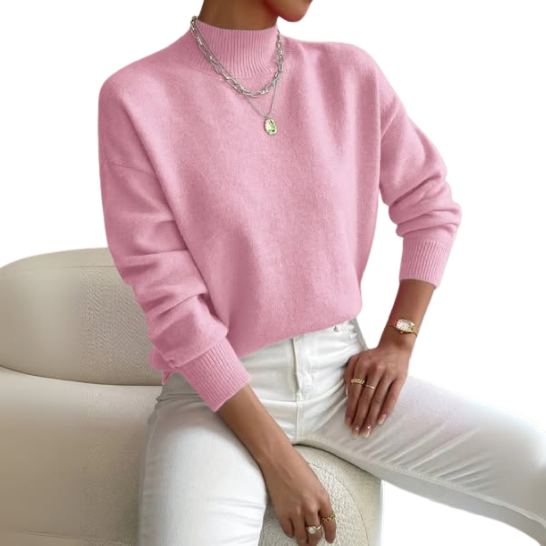 Model im rosafarbenen Pullover mit Stehkragen und Rippbündchen, kombiniert mit weißer Hose und Ketten, sitzend auf einem cremefarbenen Sessel. 1