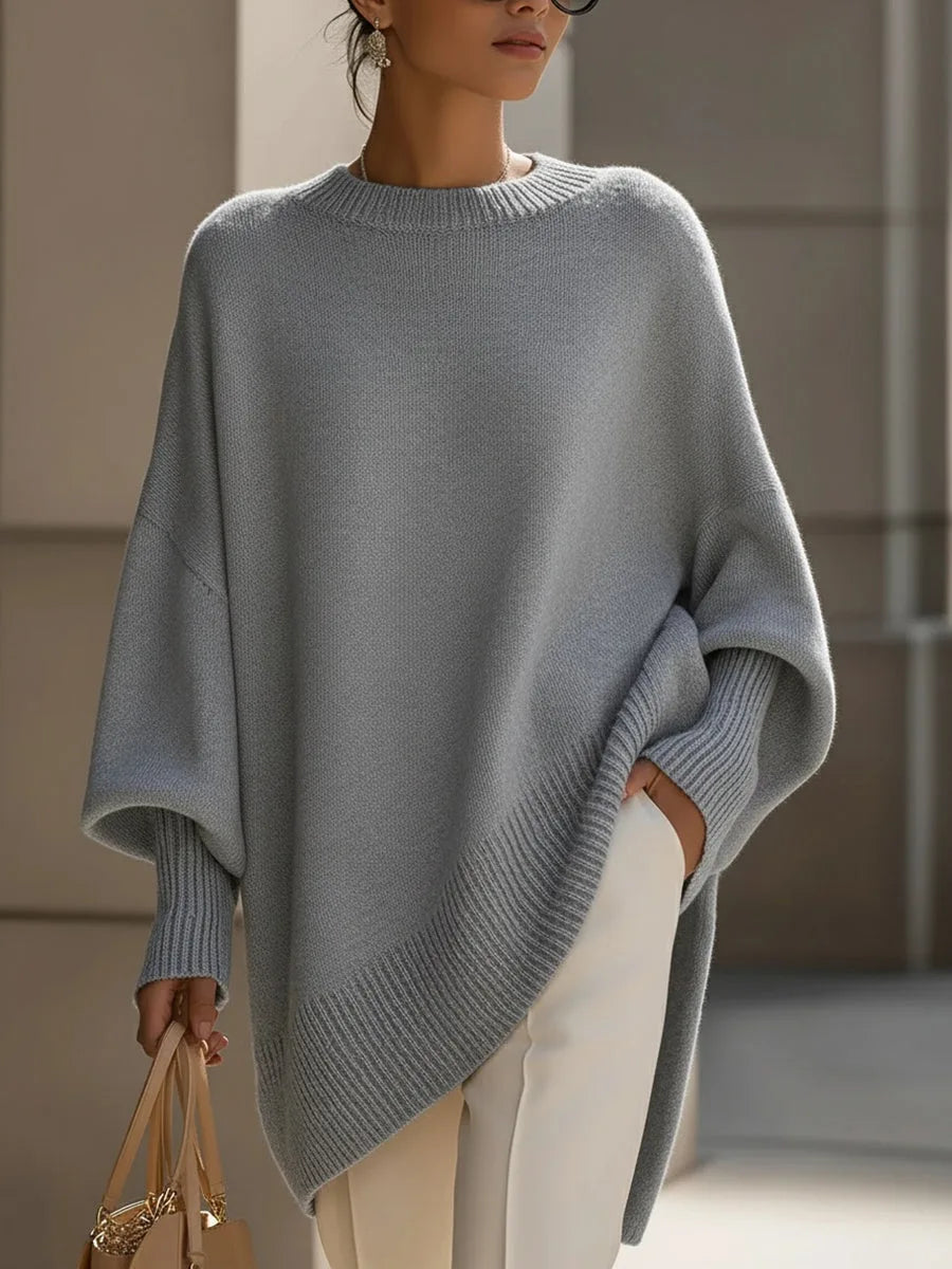 Hellgrauer Pullover mit gerippten Bündchen und schräg verlaufendem Saum, an einer Frau mit weißer Hose und Handtasche. 9