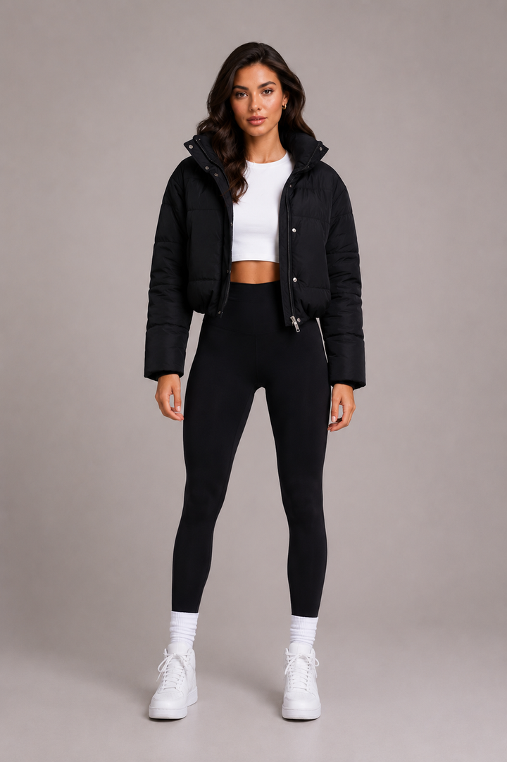 Ganzkörperansicht von vorn: Model trägt die kurze schwarze Steppjacke offen zu weißem Crop-Top, schwarzer Leggings und weißen Sneakern. 2