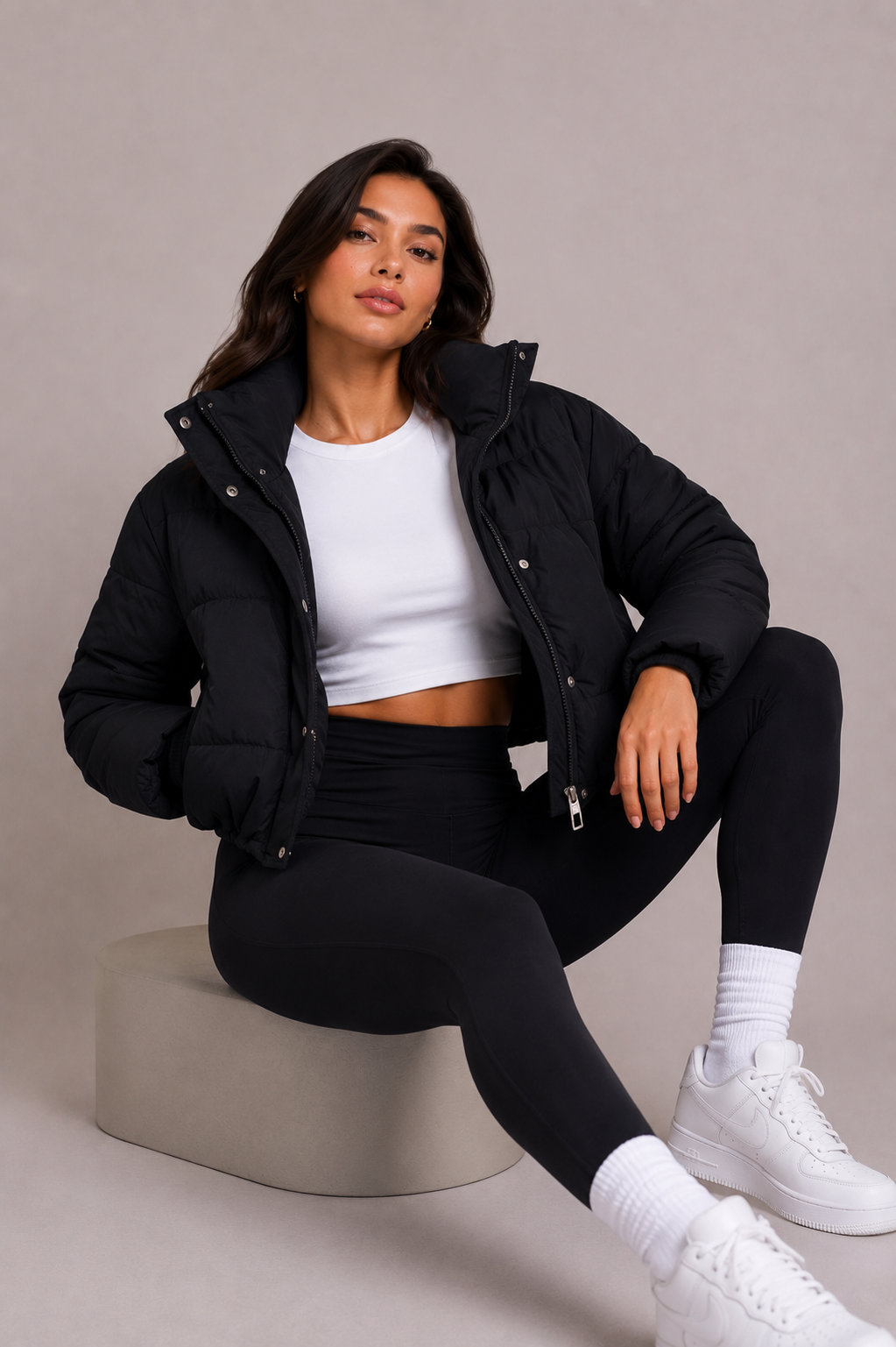 Model sitzt auf einem Podest und trägt die offene schwarze Steppjacke mit Stehkragen über weißem Crop-Top und schwarzer Leggings. 6