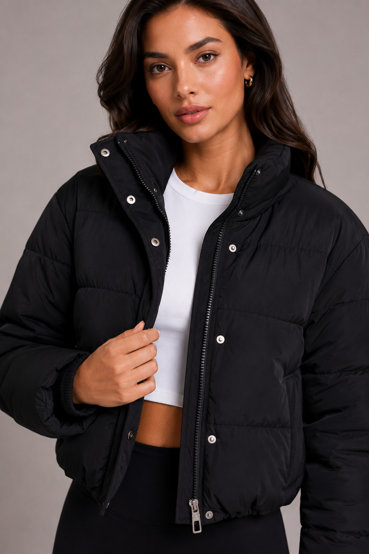 Nahaufnahme der schwarzen Steppjacke mit Stehkragen, Reißverschluss und Druckknöpfen, offen getragen über weißem Top, Model fasst den Saum. 1
