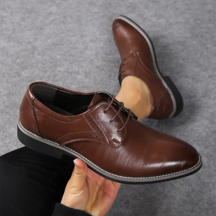 Dunkelbrauner Schnürschuh mit Zierlochung und heller Ziernaht in der Hand gehalten, daneben der zweite Schuh an einem Fuß. 2