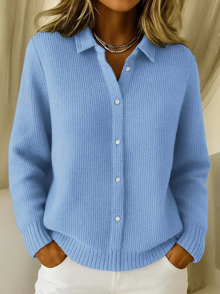 Frau trägt den strukturierten Strickpullover in Hellblau mit Kragen und Knopfleiste, die Hände in den Taschen einer weißen Hose. 1