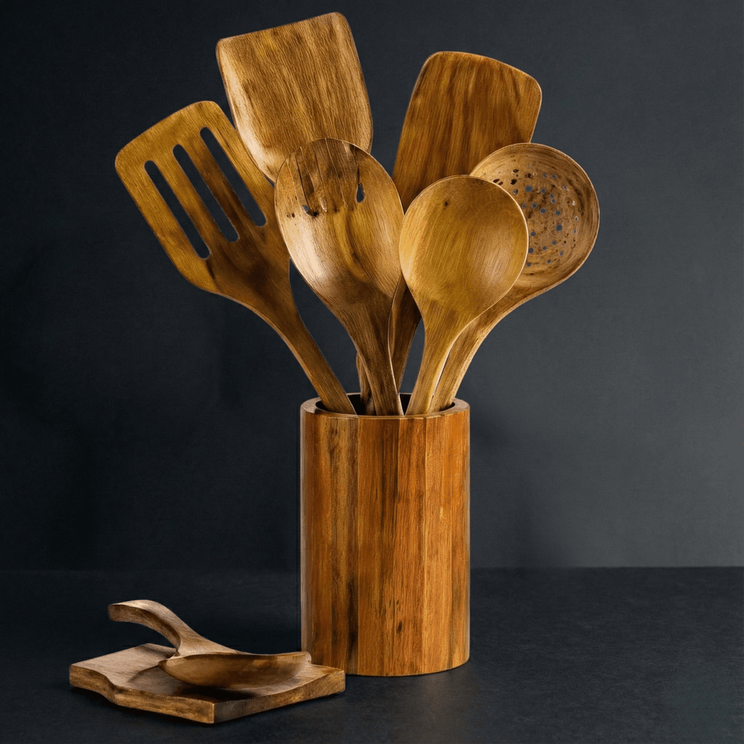 Holzutensilien in einem zylindrischen Holzbehälter vor dunklem Hintergrund, davor ein kleiner Löffelhalter aus Holz mit aufgelegtem Löffel. 1