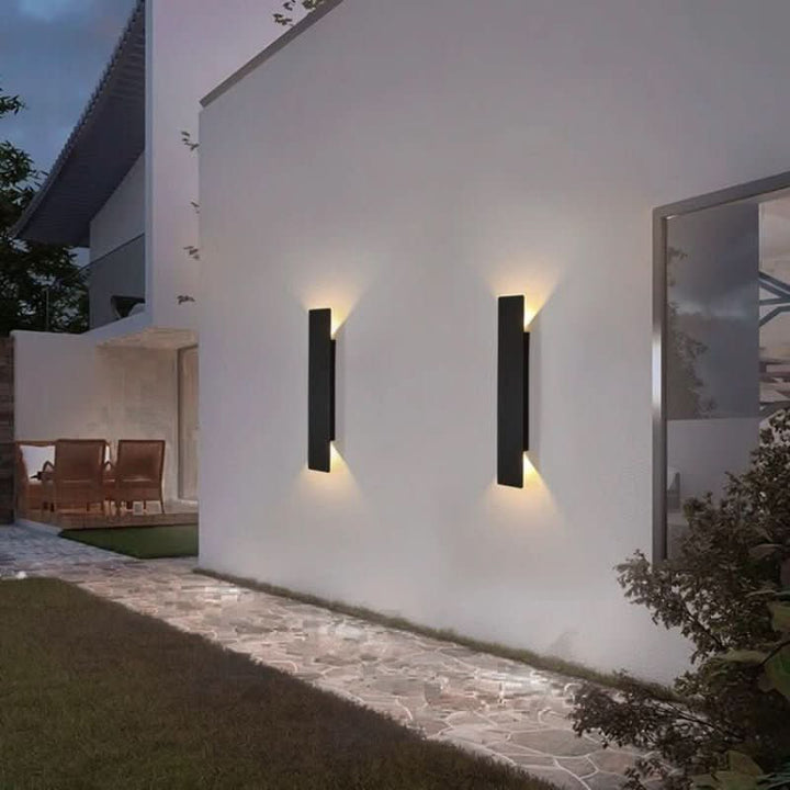 Zwei schwarze Wandleuchten nebeneinander an einer weißen Hauswand im Garten, Lichtkegel nach oben und unten, daneben ein Steinweg. 3