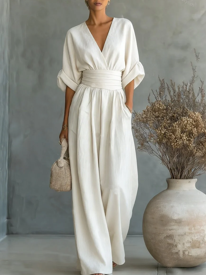 Cremeweißer Jumpsuit am Model, locker fallende Ärmel und hohe Taille, vor grauer Wand mit Vase 8