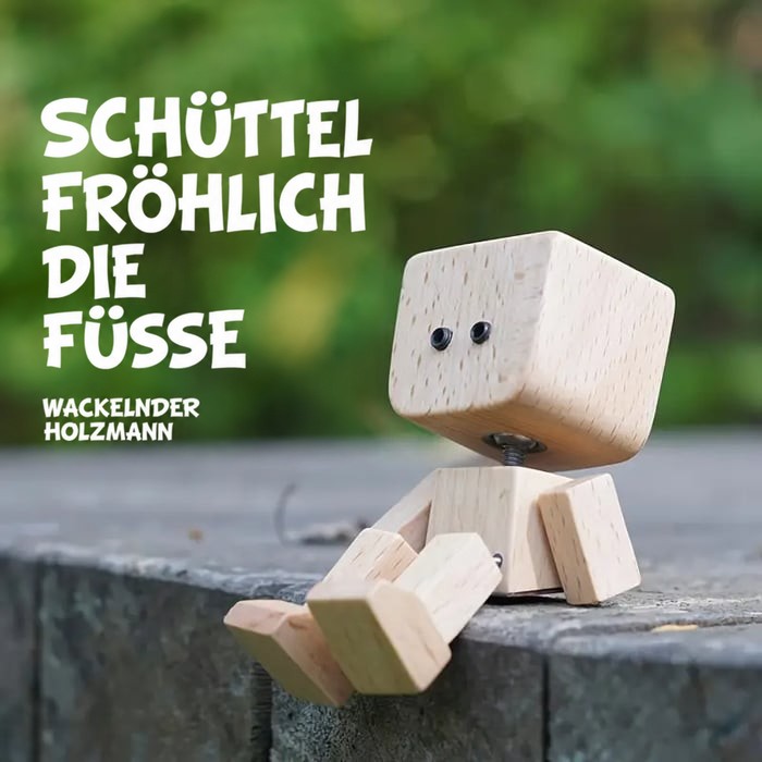 Holzfigur mit schlichten schwarzen Augen sitzt auf einer Steinkante im Grünen, die Beine baumeln locker herunter. 13