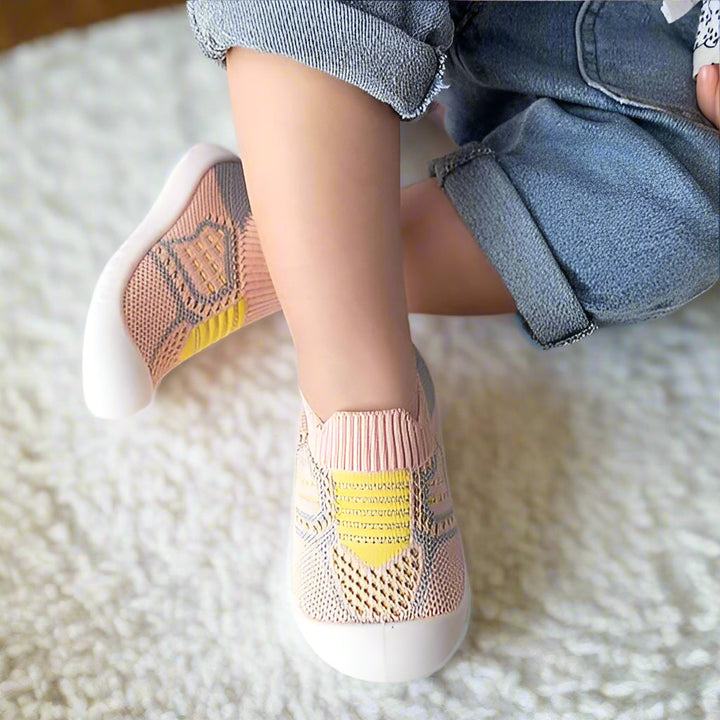Kinderfüße in rosa Strickschuhen mit gelben Streifen und Netzeinsatz auf einem weißen Teppich, ein Schuh schräg aufgestellt. 6