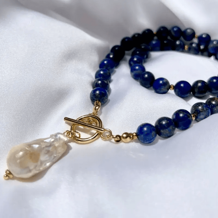 Detailaufnahme der blauen Perlen mit goldfarbenem Knebelverschluss und Barockperle auf weißem Stoff 5