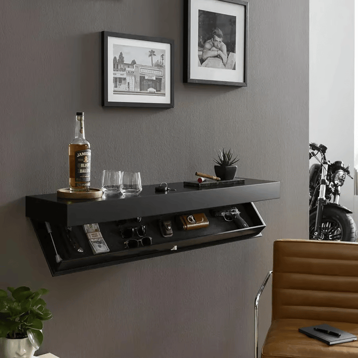 Schwarzes Wandregal an grauer Wand im Wohnraum, Fach aufgeklappt, oben Flasche, Gläser und Pflanze, daneben Bilderrahmen und Ledersessel. 4