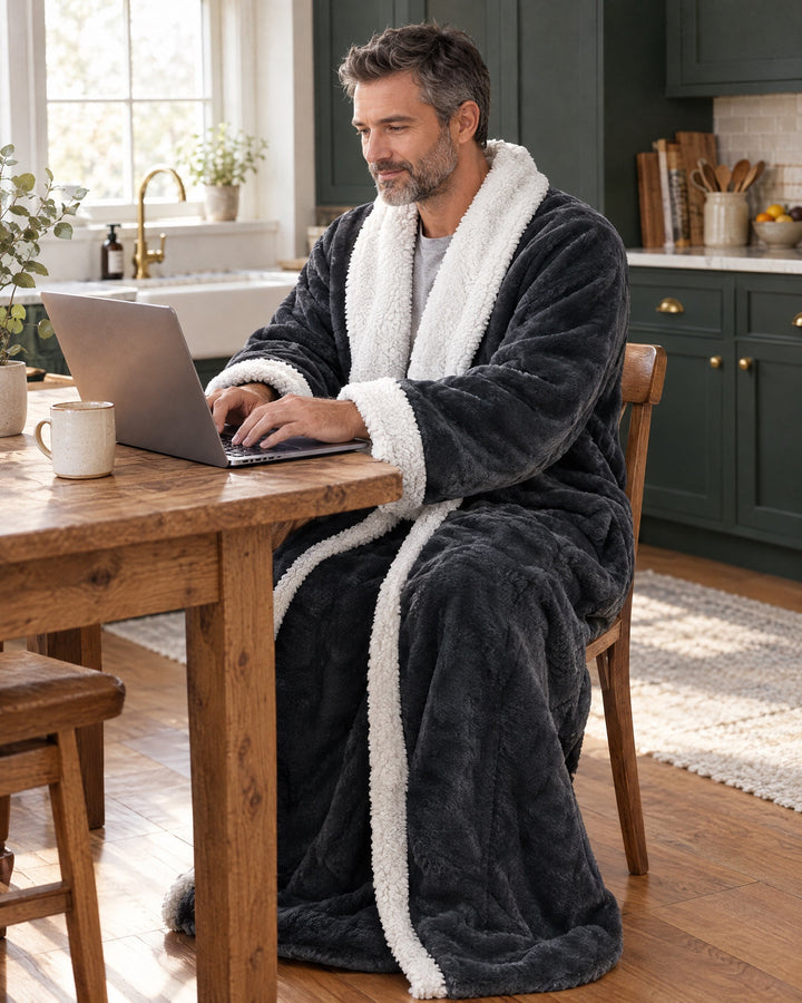 Mann sitzt im Überwurf an einem Holztisch in der Küche und arbeitet am Laptop, die Hände frei. 9
