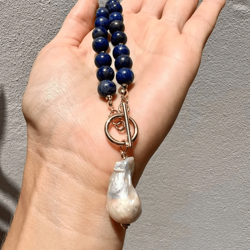 Kette mit blauen Perlen, Knebelverschluss und weißer Barockperle hängt über einer Hand vor grauer Wand 3