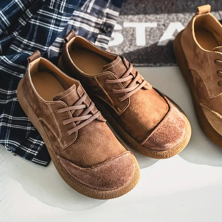 Braune Schnürschuhe in zwei Farbtönen liegen schräg nebeneinander neben einem karierten Hemd, mit Blick auf Ziernähte und Sohle. 11