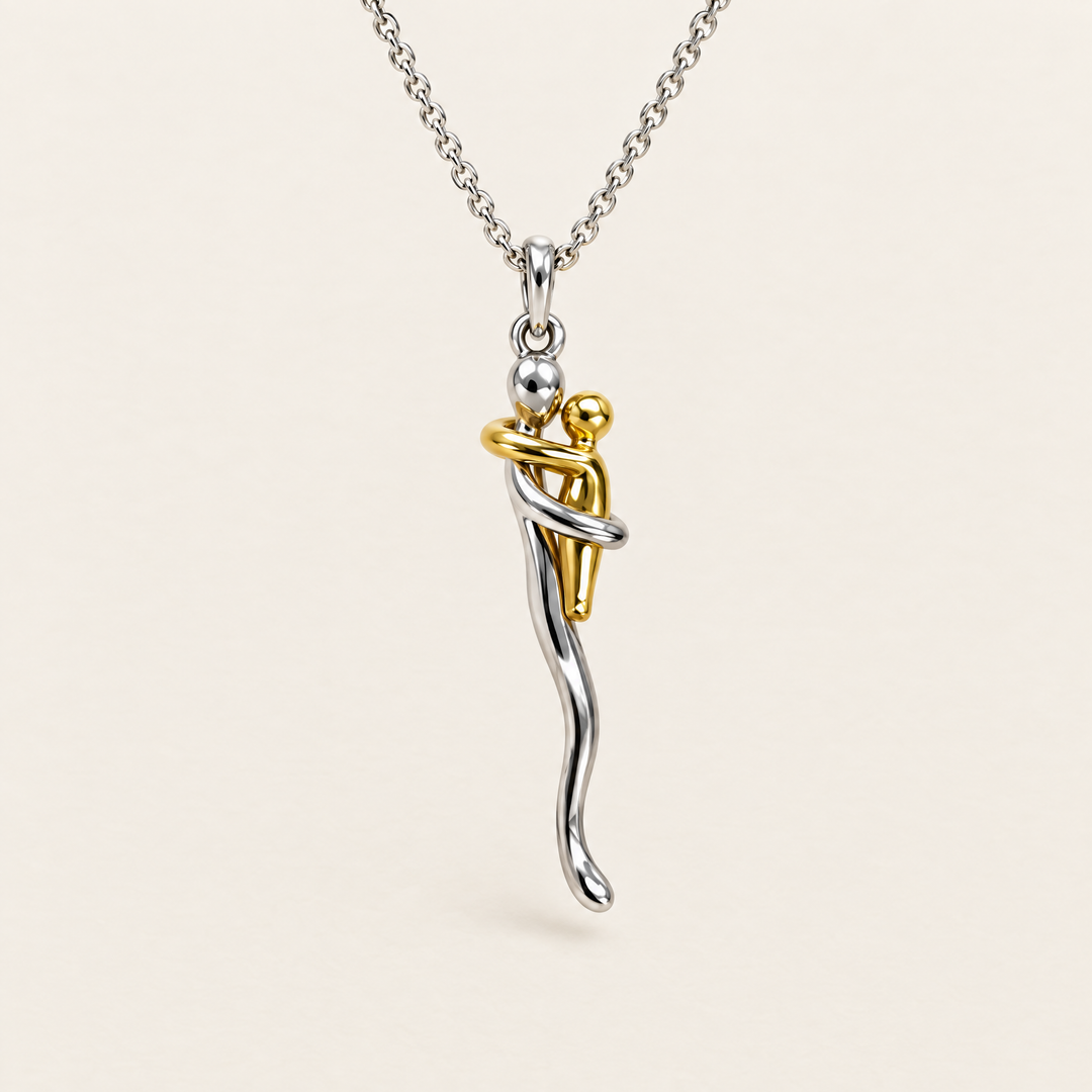 Silberfarbener Anhänger mit zwei sich umarmenden Figuren, die größere silbern, die kleinere goldfarben, an feiner Gliederkette vor hellem Hintergrund. 1